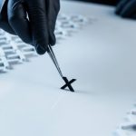 Mano con guante negro levantando pieza metálica con forma de logo de X negra con pinzas